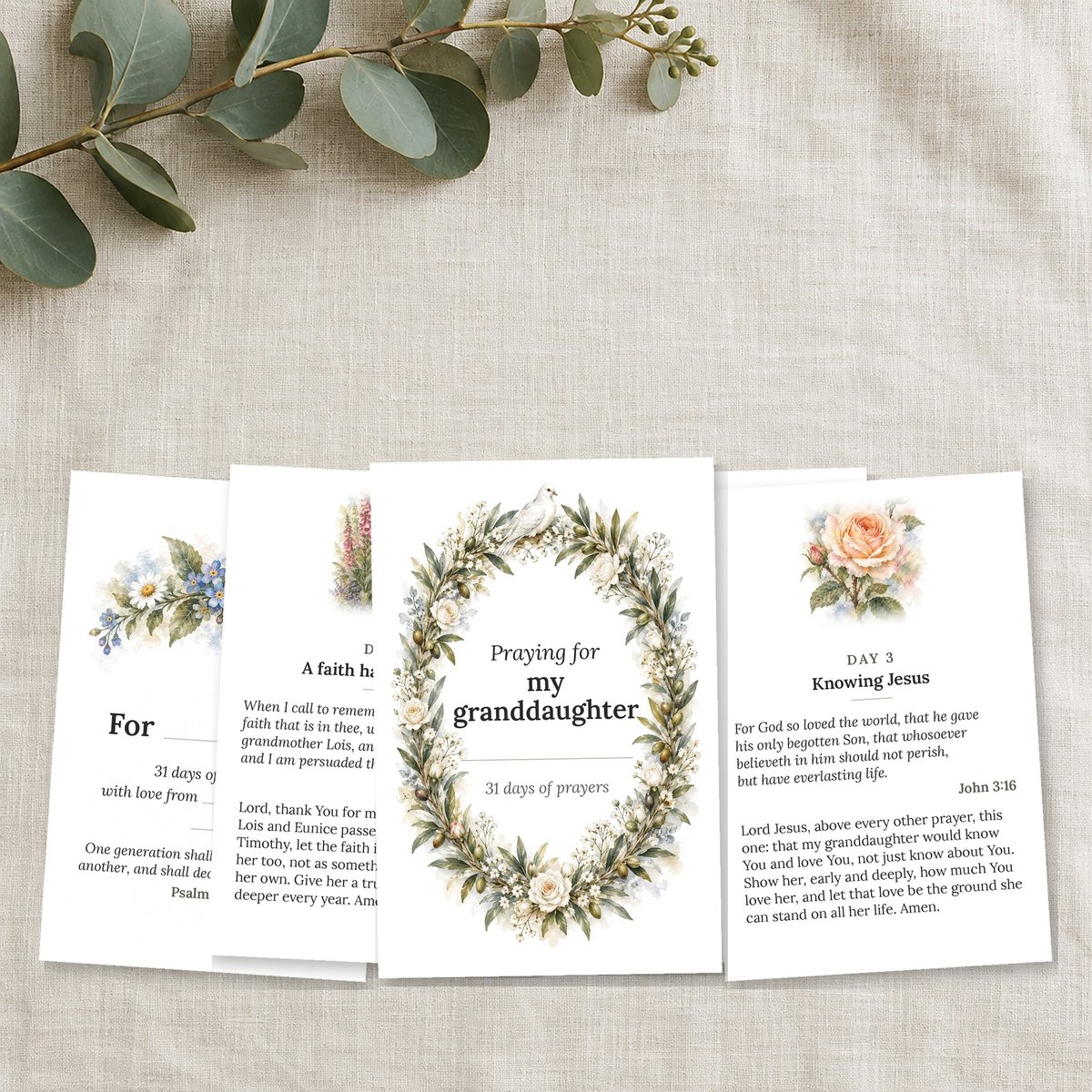 A fan of prayer cards with watercolor-style wreaths on a cream linen cloth, with a sprig of eucalyptus